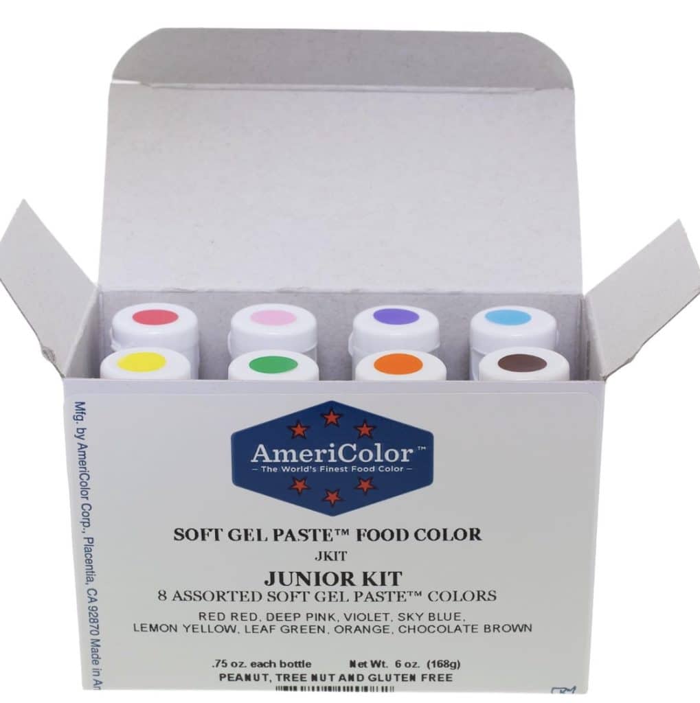 An open box showcases AmeriColor soft gel paste food colors, perfect for pink desserts and more. The set includes eight bottles with vibrant lids: red, pink, violet, blue, yellow, leaf green, orange, and chocolate. It's labeled as a peanut, tree nut, and gluten-free product.